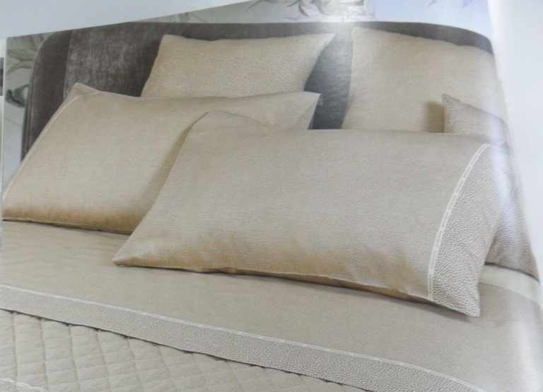 Letto con cuscini e lenzuola beige a microfantasia, bordo con fettuccia chiara, trapunta trapuntata a rombi e testiera in velluto